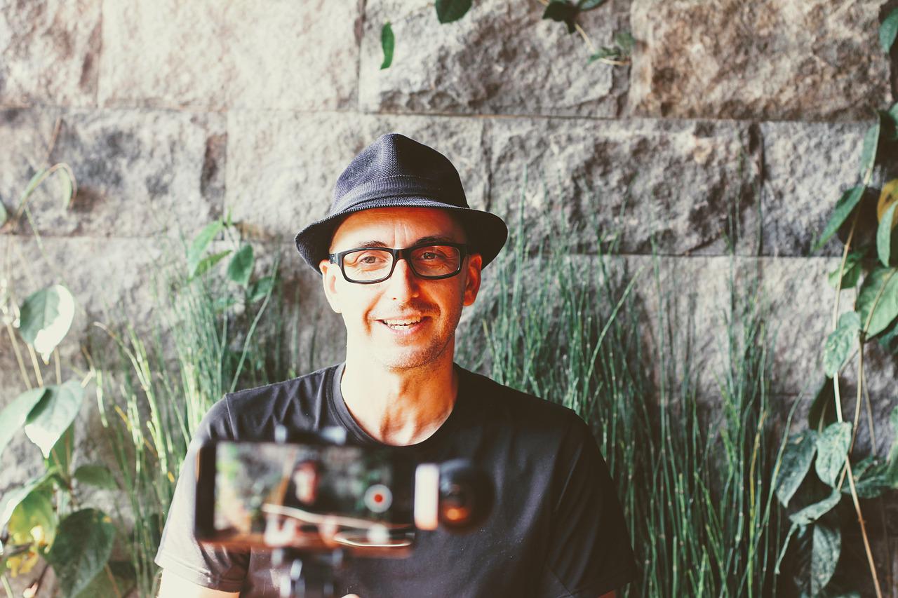 Smartphone on tripod recording a man with glasses and hat sitting outside in front of a wall with greenery